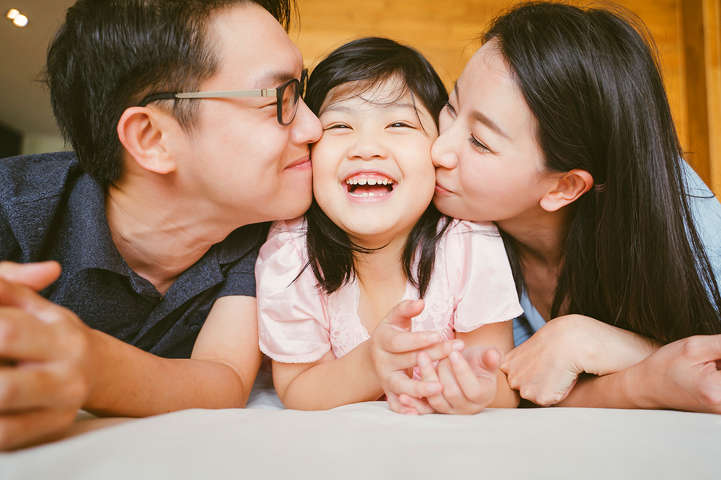 Two parents with their smiling child in between while kissing her on her cheeks.