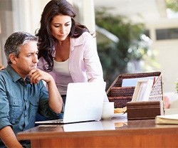 A woman standing and man sitting at a desk reviewing content on a laptop in a home office.